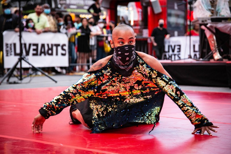 Jasmine Hearn wears a black bandana as a mask and a billowy, sequined shirt. She kneels on a red floor as pedestrians look on.